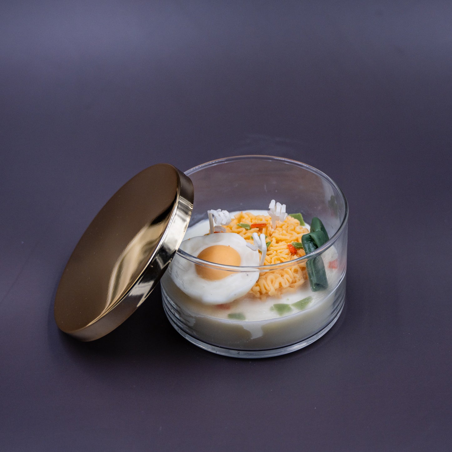 This highly realistic, handcrafted candle is a culinary work of art, designed to perfectly mimic a bowl of ramen noodles with a poached egg, kelp knot, carrots, and other vegetable toppings, all in a white savory bone broth base. Primarily made from a blend of natural soy and beeswax, this unique item is an ideal novelty decor piece. The entire assembly is presented in a glass jar with rose golden lid.