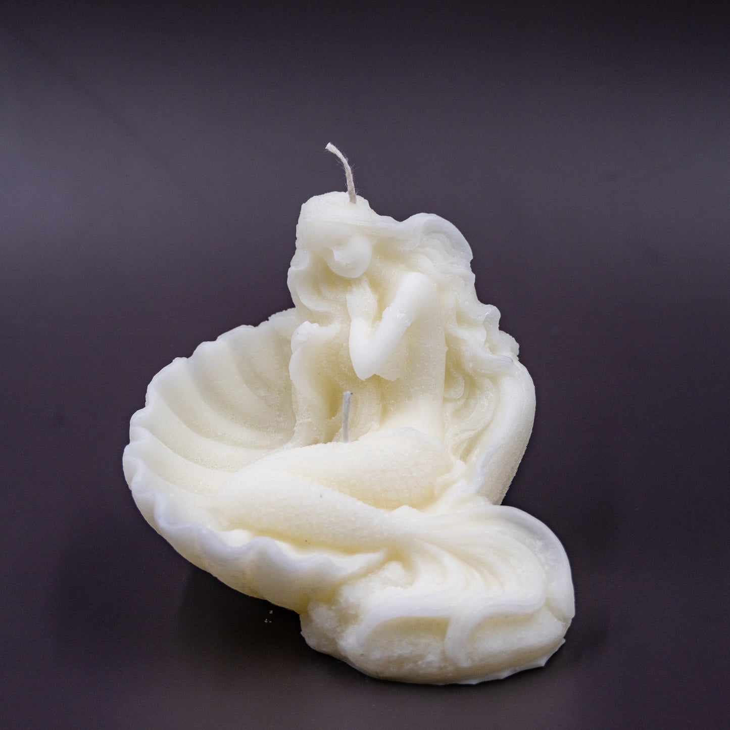 The candle features an intricate, whimsical sculpture of a serene mermaid nestled in or wrapped around a spiral seashell, designed to add a serene, romantic, storybook or coastal charm to any space.