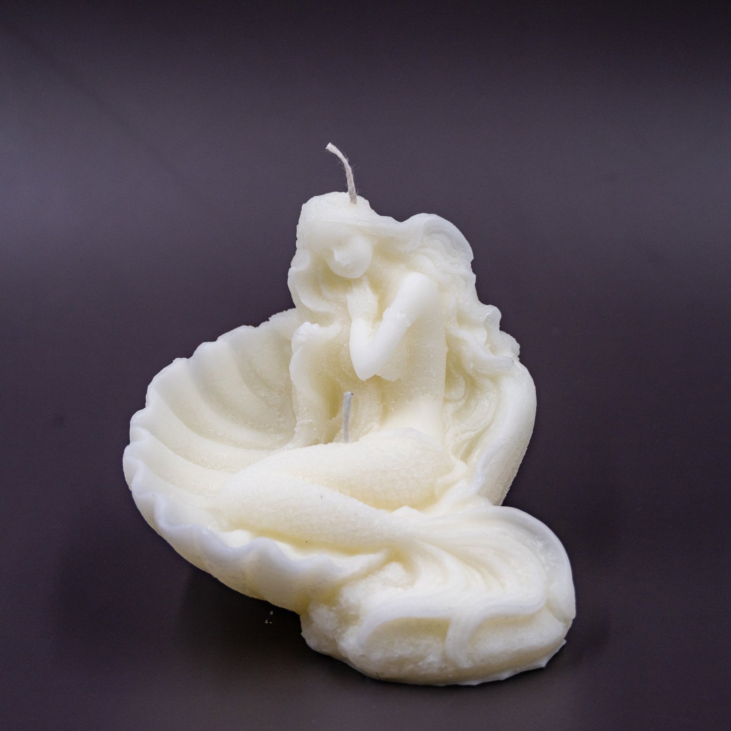 The candle features an intricate, whimsical sculpture of a serene mermaid nestled in or wrapped around a spiral seashell, designed to add a serene, romantic, storybook or coastal charm to any space.