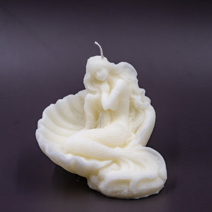 The candle features an intricate, whimsical sculpture of a serene mermaid nestled in or wrapped around a spiral seashell, designed to add a serene, romantic, storybook or coastal charm to any space.