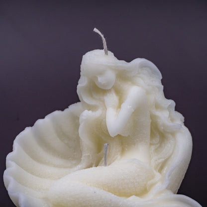 The candle features an intricate, whimsical sculpture of a serene mermaid nestled in or wrapped around a spiral seashell, designed to add a serene, romantic, storybook or coastal charm to any space.