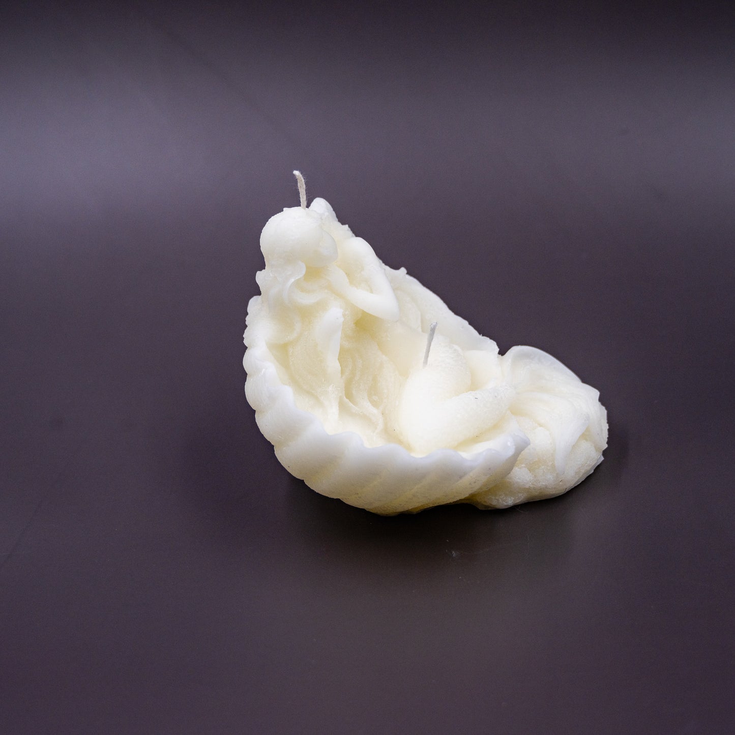 The candle features an intricate, whimsical sculpture of a serene mermaid nestled in or wrapped around a spiral seashell, designed to add a serene, romantic, storybook or coastal charm to any space.