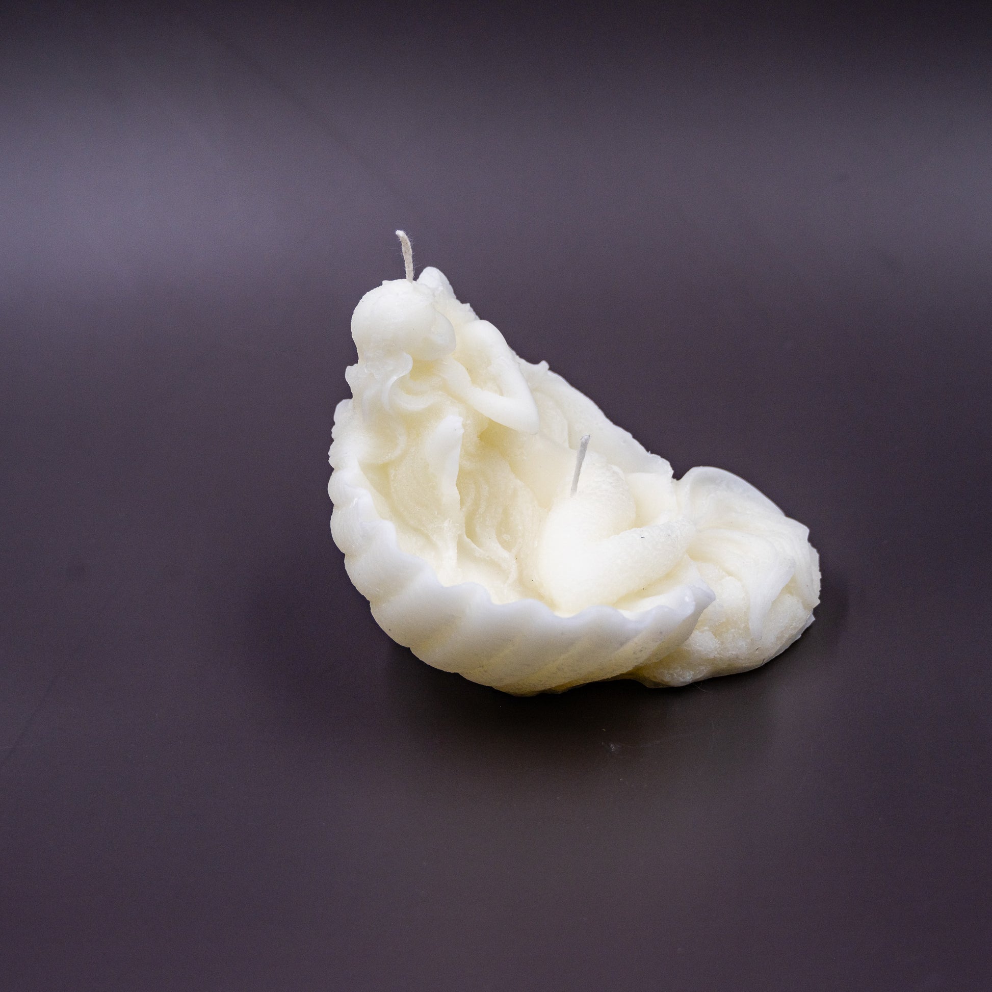 The candle features an intricate, whimsical sculpture of a serene mermaid nestled in or wrapped around a spiral seashell, designed to add a serene, romantic, storybook or coastal charm to any space.