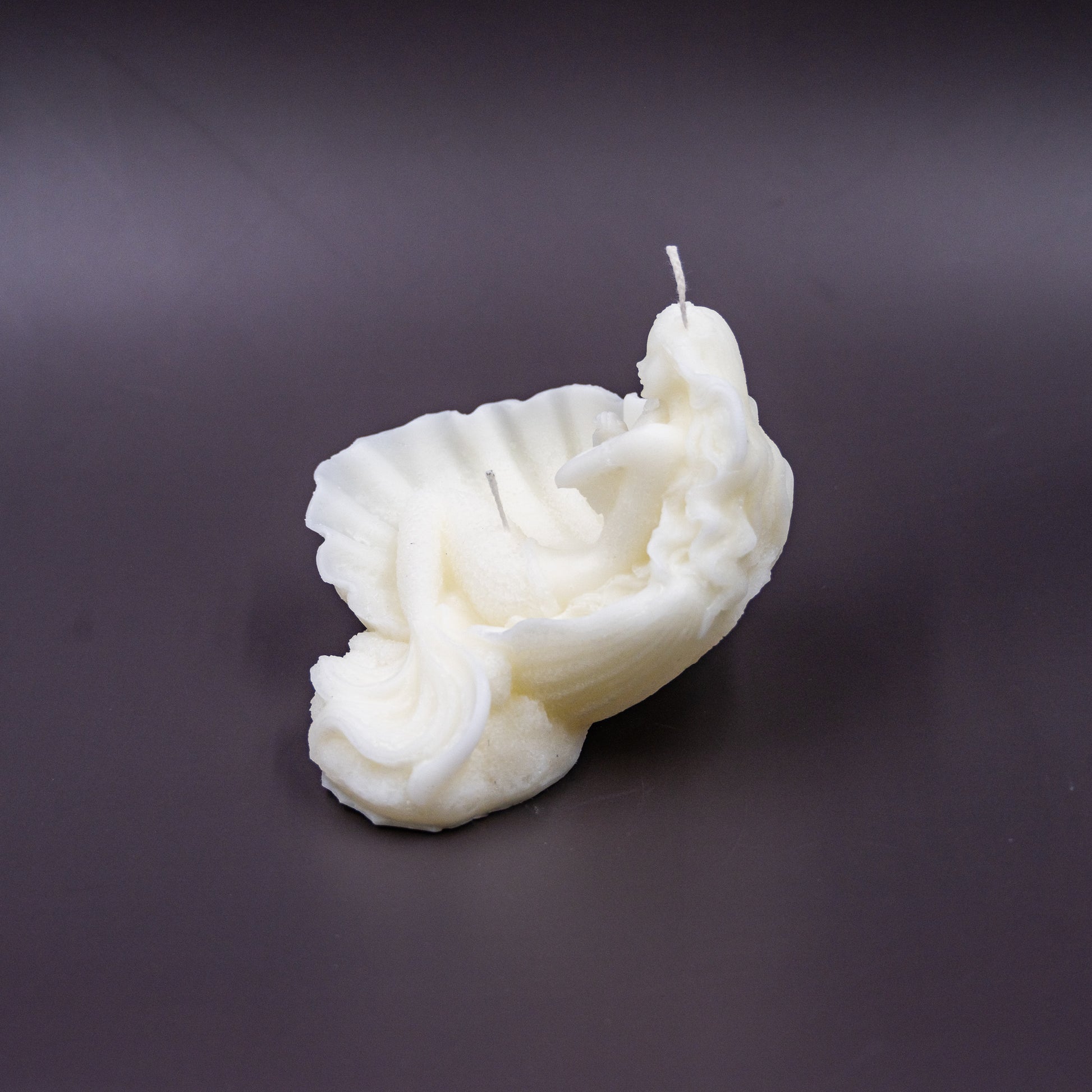 The candle features an intricate, whimsical sculpture of a serene mermaid nestled in or wrapped around a spiral seashell, designed to add a serene, romantic, storybook or coastal charm to any space.