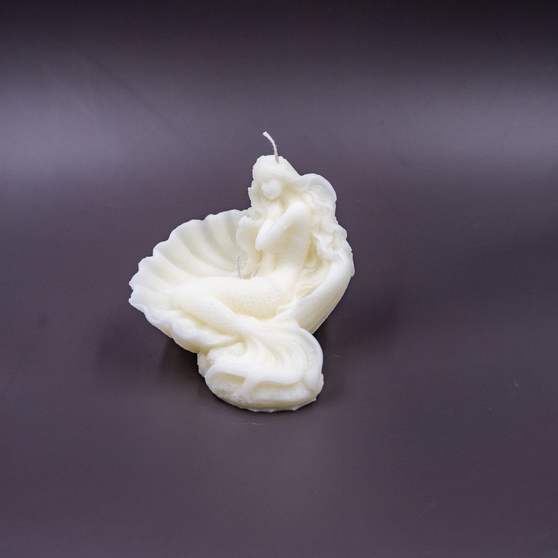 The candle features an intricate, whimsical sculpture of a serene mermaid nestled in or wrapped around a spiral seashell, designed to add a serene, romantic, storybook or coastal charm to any space.