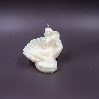 The candle features an intricate, whimsical sculpture of a serene mermaid nestled in or wrapped around a spiral seashell, designed to add a serene, romantic, storybook or coastal charm to any space.