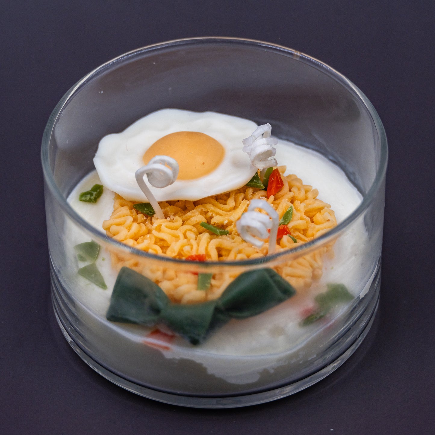 This highly realistic, handcrafted candle is a culinary work of art, designed to perfectly mimic a bowl of ramen noodles with a poached egg, kelp knot, carrots, and other vegetable toppings, all in a white savory bone broth base. Primarily made from a blend of natural soy and beeswax, this unique item is an ideal novelty decor piece. The entire assembly is presented in a glass jar with rose golden lid.