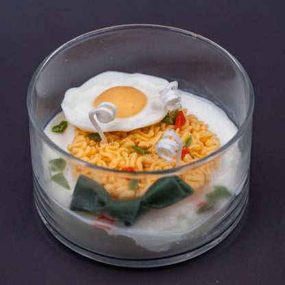 This highly realistic, handcrafted candle is a culinary work of art, designed to perfectly mimic a bowl of ramen noodles with a poached egg, kelp knot, carrots, and other vegetable toppings, all in a white savory bone broth base. Primarily made from a blend of natural soy and beeswax, this unique item is an ideal novelty decor piece. The entire assembly is presented in a glass jar with rose golden lid.