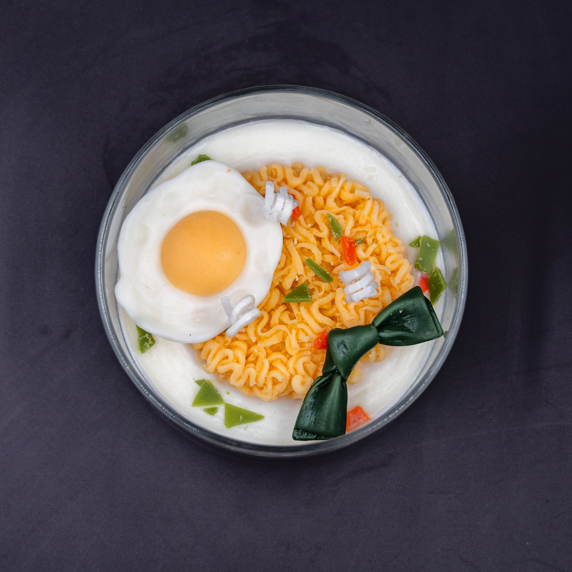 This highly realistic, handcrafted candle is a culinary work of art, designed to perfectly mimic a bowl of ramen noodles with a poached egg, kelp knot, carrots, and other vegetable toppings, all in a white savory bone broth base. Primarily made from a blend of natural soy and beeswax, this unique item is an ideal novelty decor piece. The entire assembly is presented in a glass jar with rose golden lid.