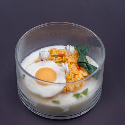This highly realistic, handcrafted candle is a culinary work of art, designed to perfectly mimic a bowl of ramen noodles with a poached egg, kelp knot, carrots, and other vegetable toppings, all in a white savory bone broth base. Primarily made from a blend of natural soy and beeswax, this unique item is an ideal novelty decor piece. The entire assembly is presented in a glass jar with rose golden lid.