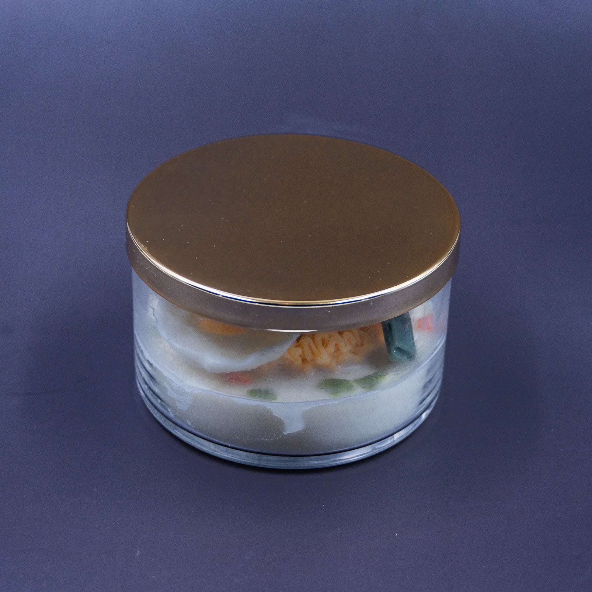 This highly realistic, handcrafted candle is a culinary work of art, designed to perfectly mimic a bowl of ramen noodles with a poached egg, kelp knot, carrots, and other vegetable toppings, all in a white savory bone broth base. Primarily made from a blend of natural soy and beeswax, this unique item is an ideal novelty decor piece. The entire assembly is presented in a glass jar with rose golden lid.
